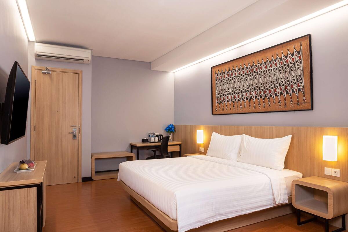 Ein Hotelzimmer mit einem Bett, Nachttischen, einem Schreibtisch, einem Flachbildfernseher und einem an der Wand befestigten Kunstwerk.