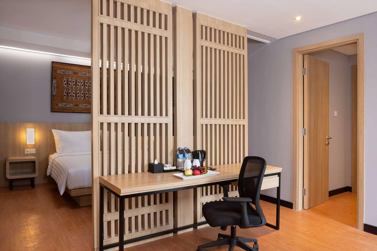 Ein Hotelzimmer mit Holztisch, einem Bett, einem Nachttisch, einem Sessel und einem an der Wand hängenden Kunstwerk.