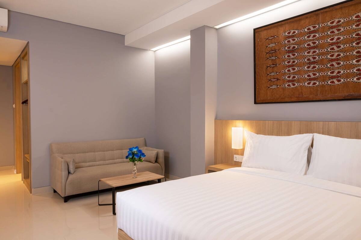Ein modernes Hotelzimmer mit einem großen Bett, einer Sitzecke, einem kleinen Tisch mit Blumenvase und einem Wandbild.