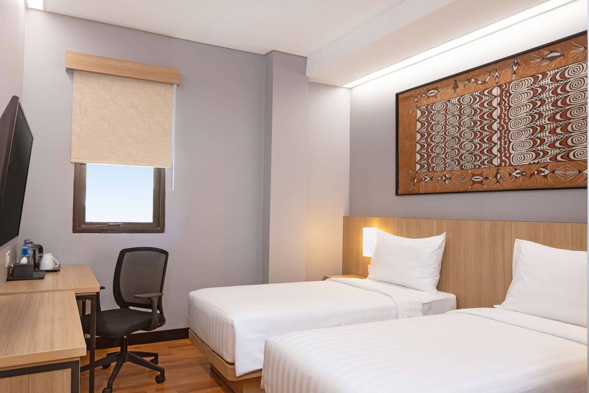 Ein Hotelzimmer mit zwei Betten, einem Schreibtisch mit Stuhl, einem Fernseher und einem Fenster mit Jalousie.