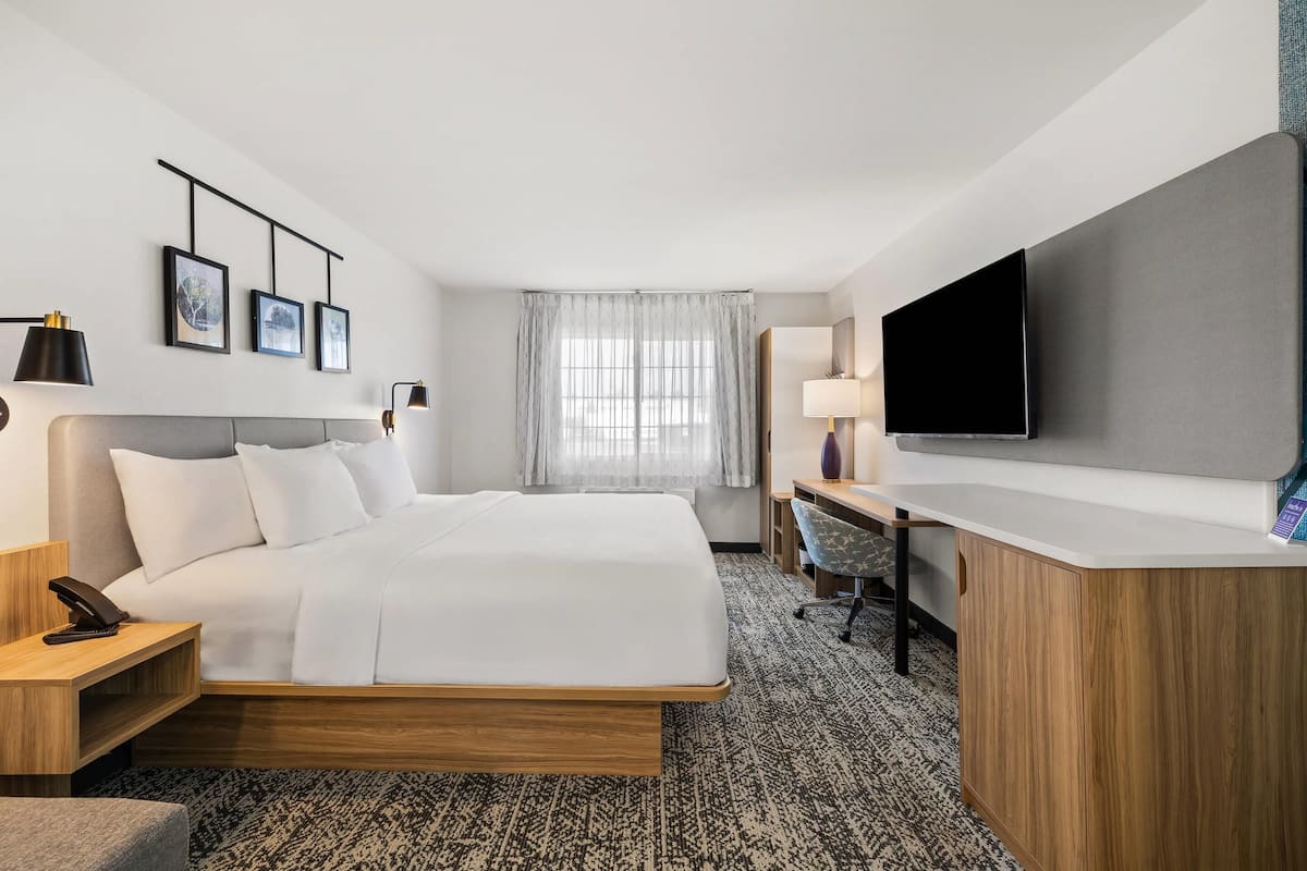 A hotel room with a bed, desk, TV, and artwork on the wall.