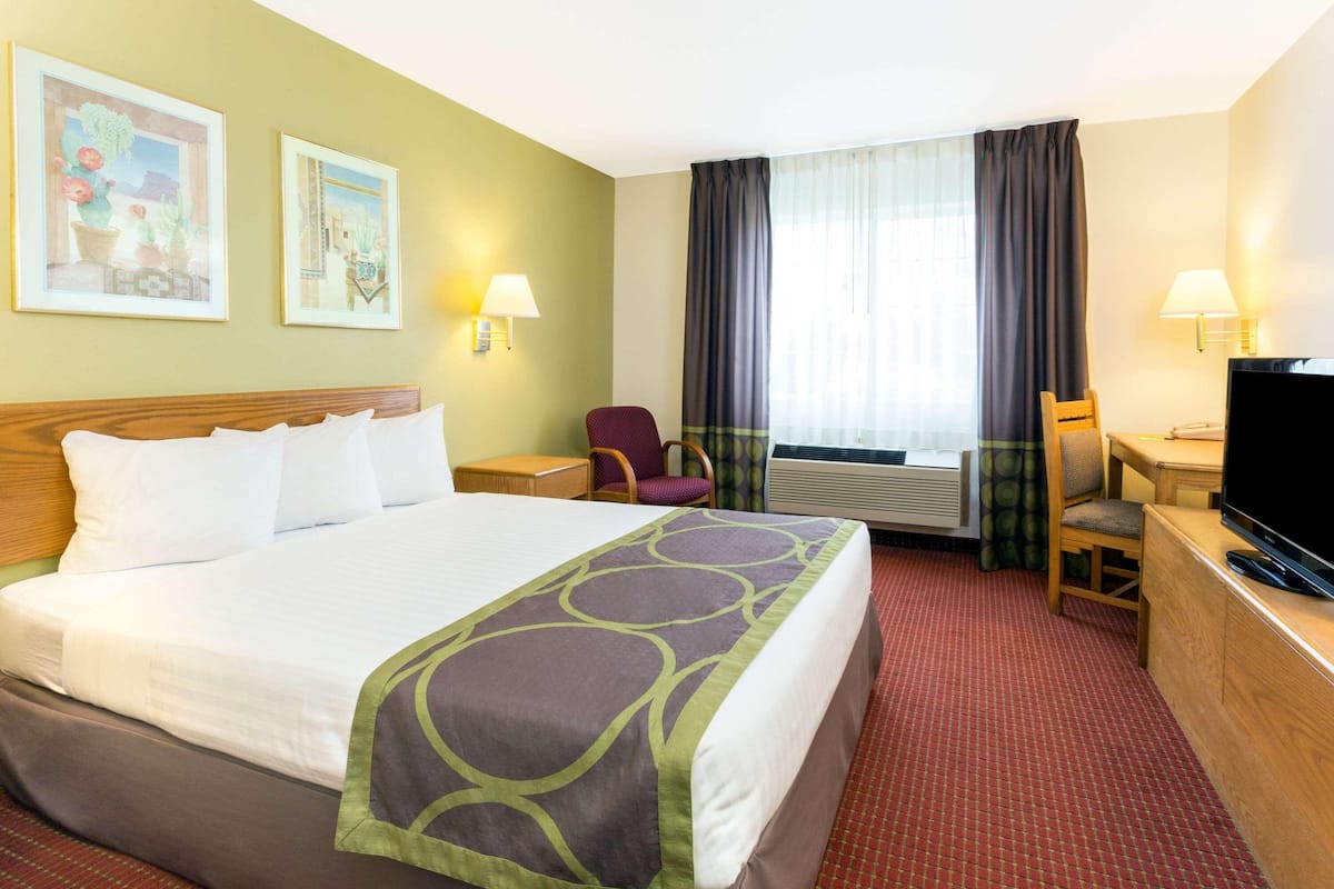 A hotel room with a large bed, two wall-mounted lamps, a desk with a television, and two windows with curtains.