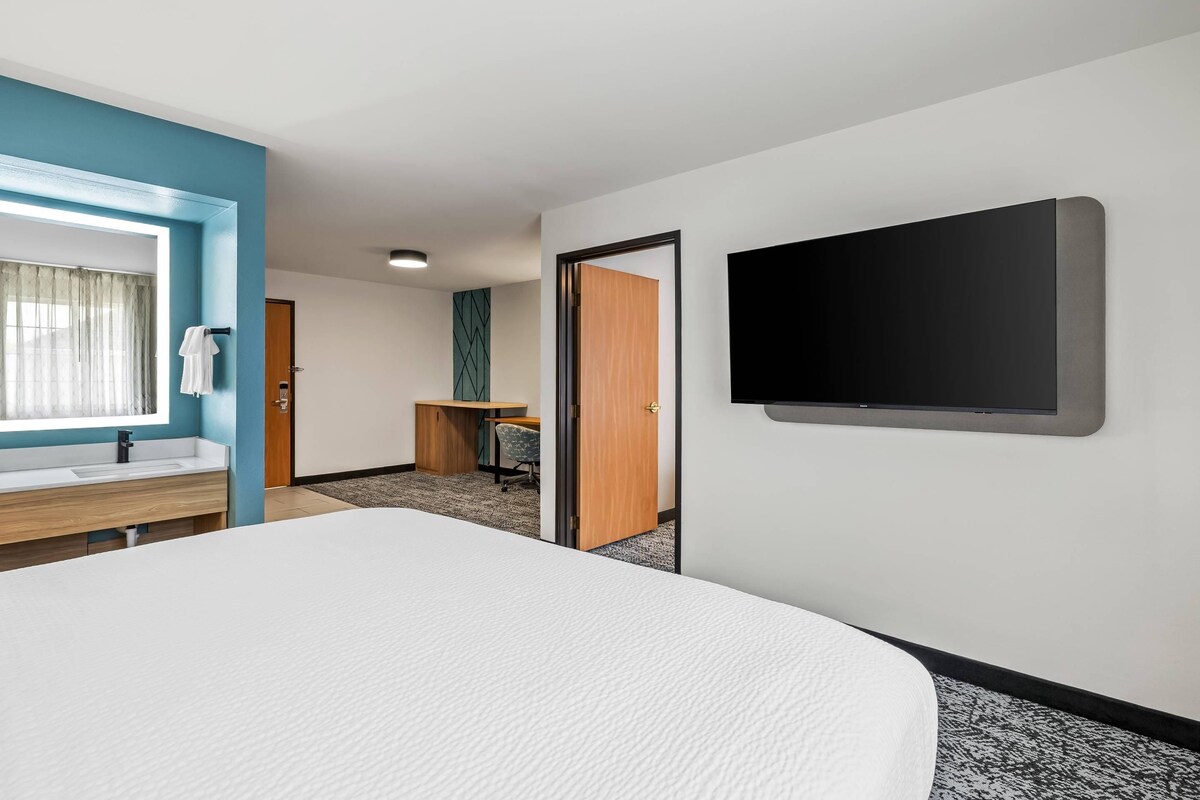 Habitación de hotel con cama, escritorio, televisión, baño con lavamanos y puerta que da a otra habitación.
