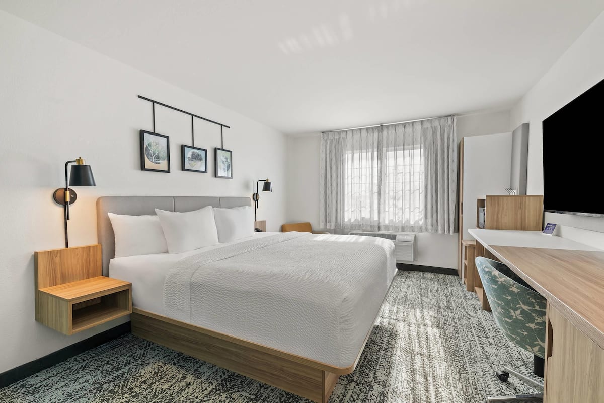A hotel room with a bed, bedside tables, a desk, a chair, a TV, and a window with curtains.
