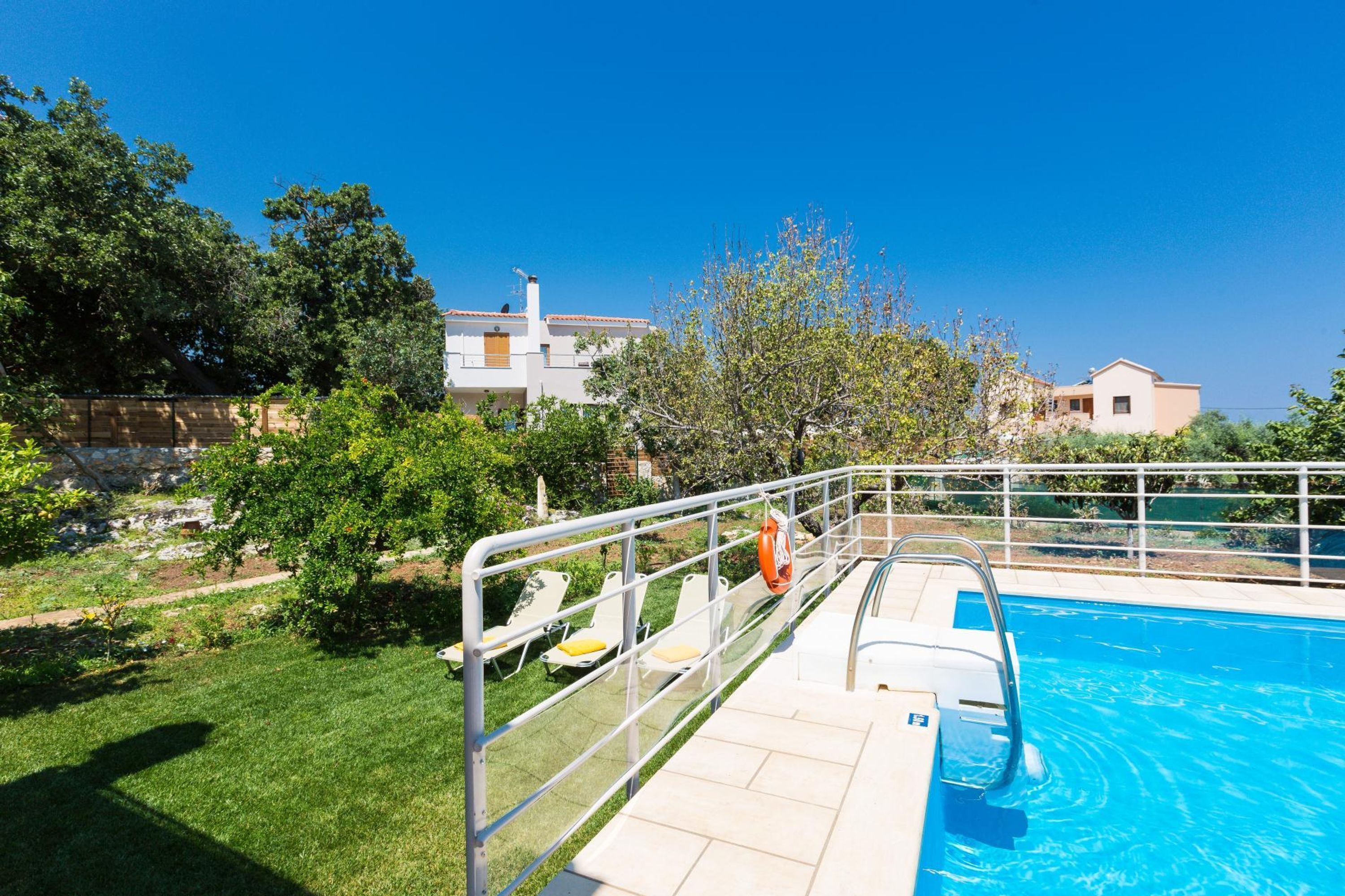 A swimming pool with a metal deck and lounge chairs, surrounded by greenery and buildings.