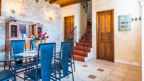 A dining area with a glass table and chairs, a stone wall, wooden ceiling beams, and a staircase with wooden doors. - Villa Marcos Chara in Asteri (Rethymno)