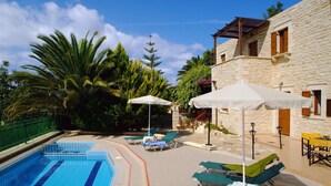A swimming pool with lounge chairs and umbrellas, surrounded by stone buildings and greenery. - Villa Marcos Chara in Asteri (Rethymno)