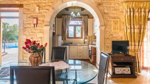 A stone-walled room with a glass dining table, chairs, a TV, and a kitchen area visible through an archway. - Villa Marcos Chara in Asteri (Rethymno)
