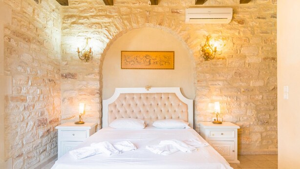 A bedroom with a stone wall, a tufted headboard, two bedside tables with lamps, and a bed with white linens. - Villa Marcos Chara in Asteri (Rethymno)