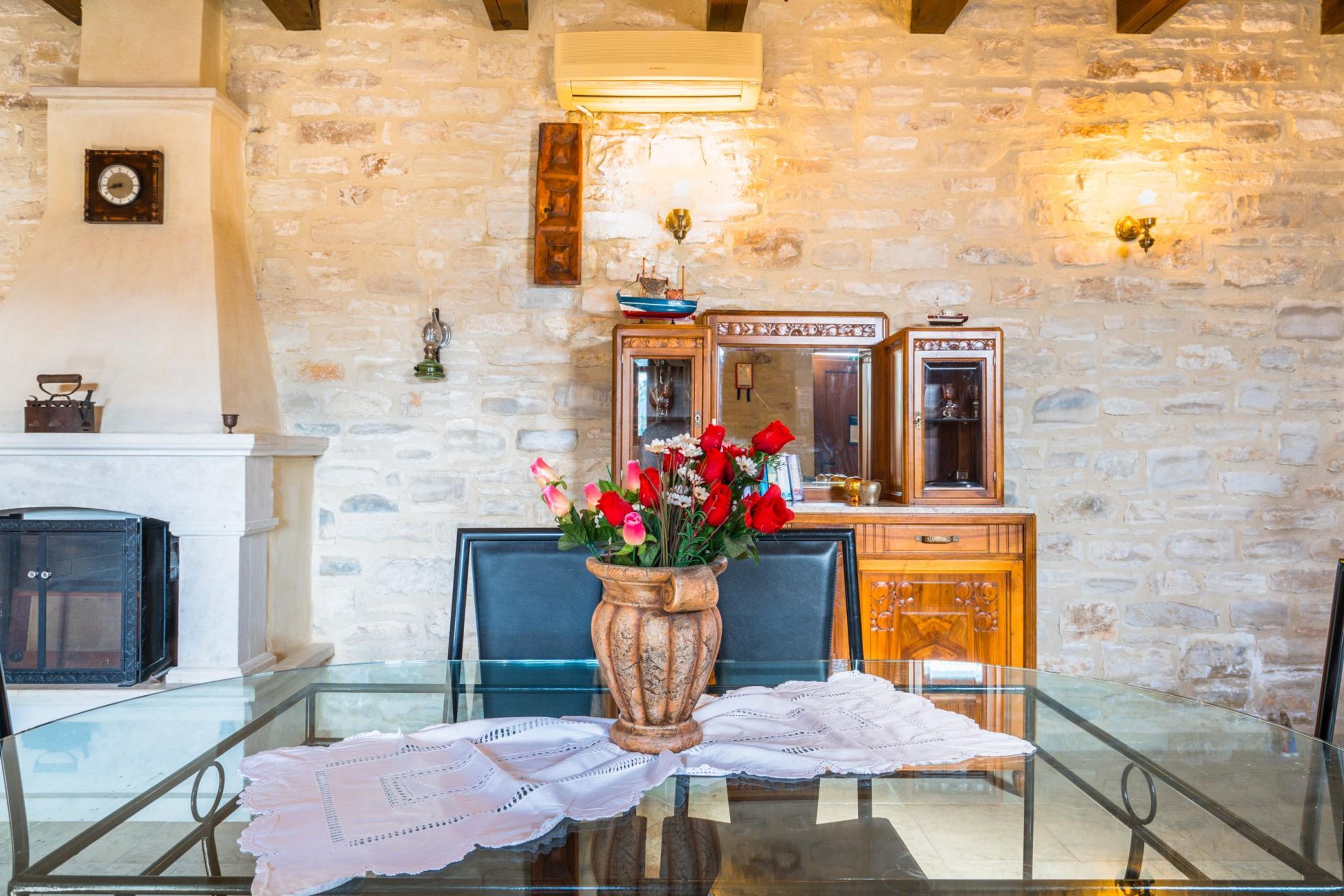 A dining area with a glass table, chairs, a vase with flowers, a wooden cabinet, and a stone wall.