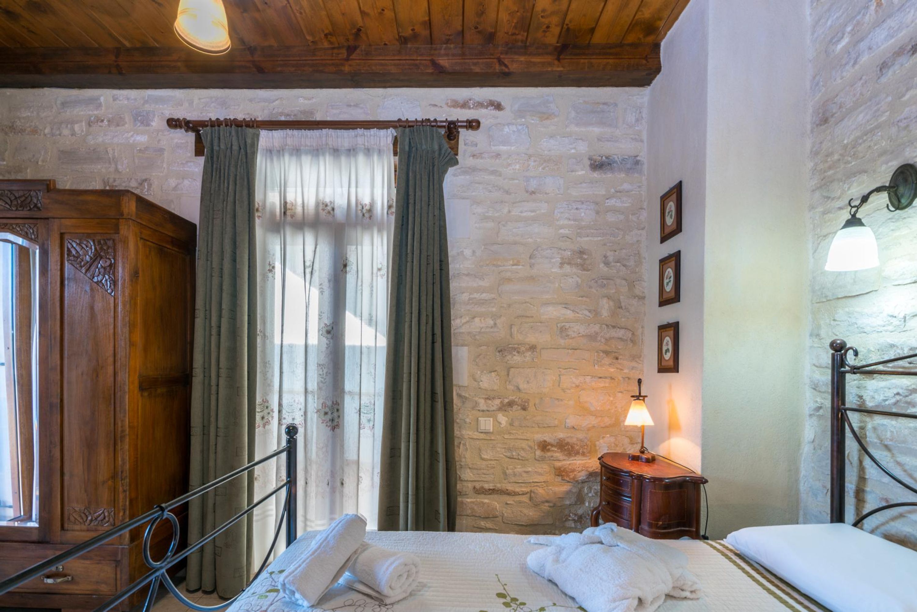 A bedroom with a wooden bed, a nightstand with a lamp, a window with curtains, and a stone wall.