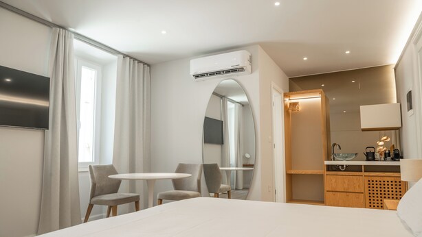 A modern hotel room with a large bed, two chairs, a small table, a mirror, a kitchenette, and an air conditioning unit.