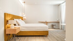 A modern bedroom with a wooden headboard, a bedside table with a lamp, a small round stool, and a view of the room through a window.