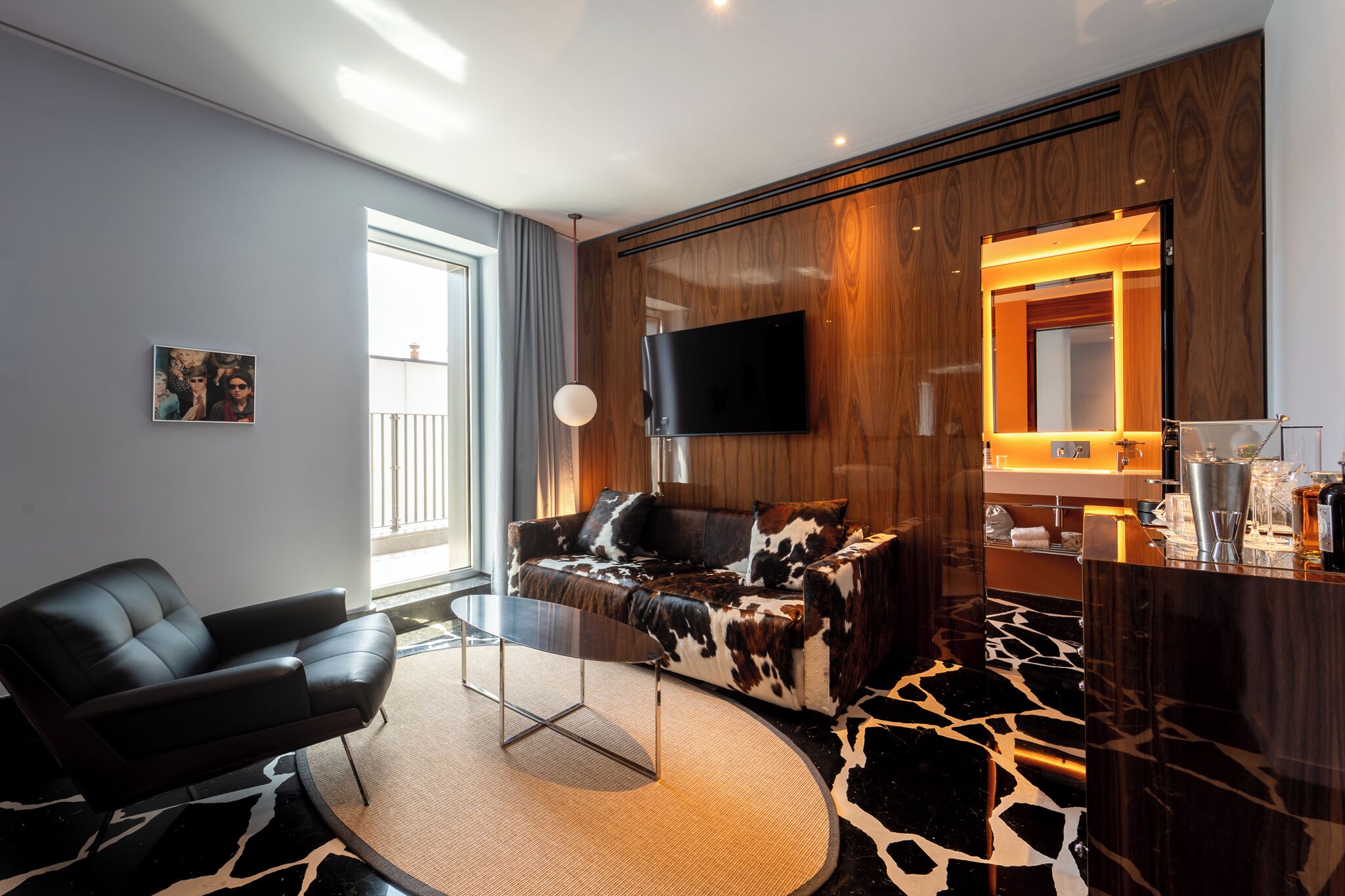 Un salon moderne avec un canapé en cuir, une table basse en verre, un téléviseur à écran plat, un mur d’accent en bois et un coin cuisine équipé d’un évier et d’un plan de travail.
