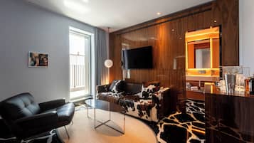 Un salon moderne avec un canapé en cuir, une table basse en verre, un téléviseur à écran plat, un mur d’accent en bois et un coin cuisine équipé d’un évier et d’un plan de travail.
