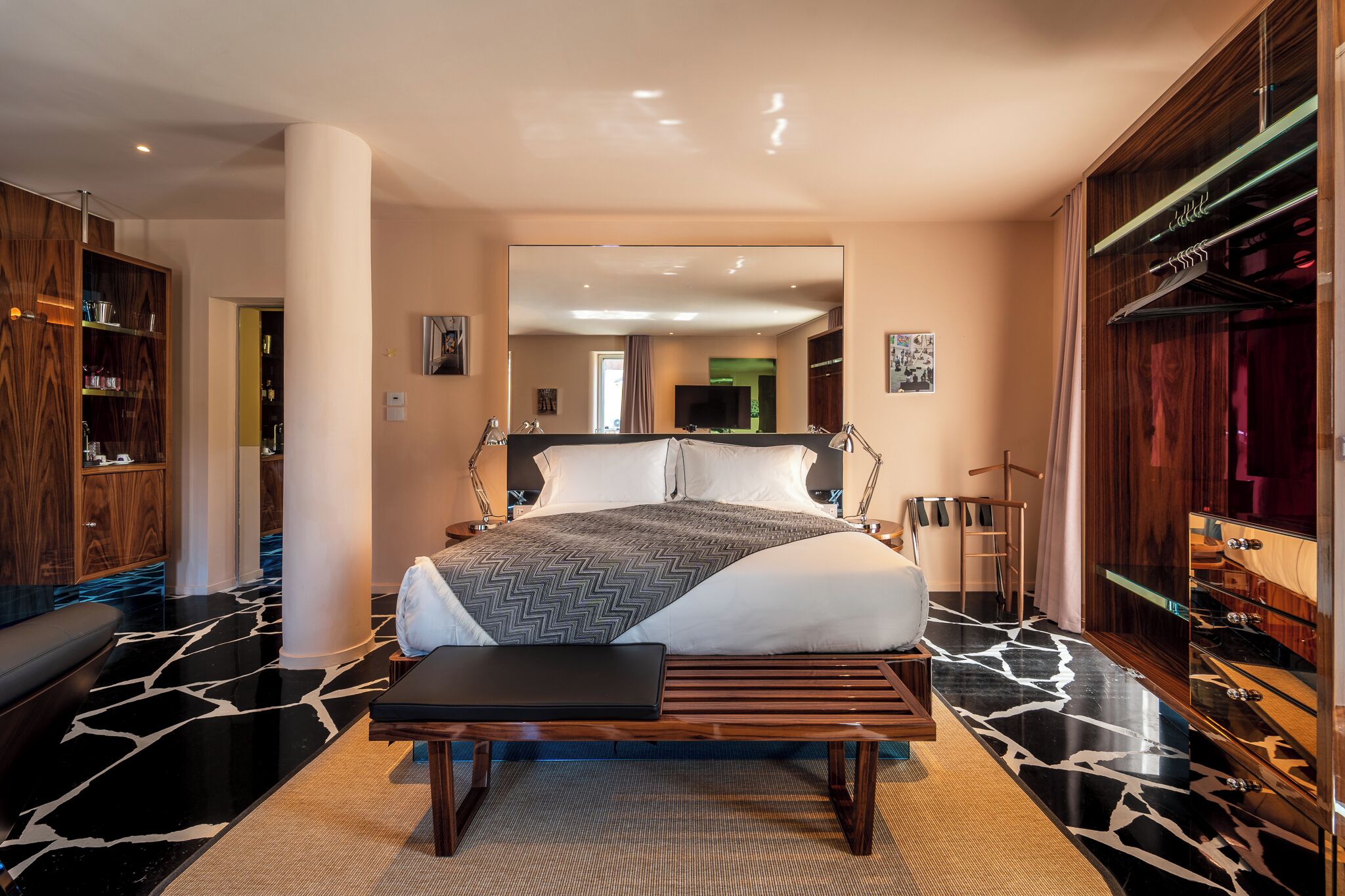 A modern bedroom with a large bed, a bench, a wardrobe, and a mirror.