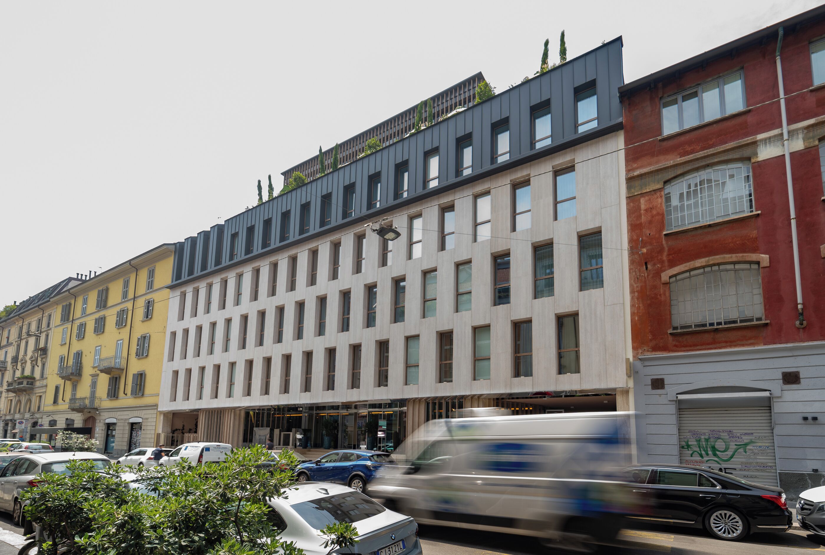 A multi-story building with a modern facade, parked cars, and a moving vehicle.