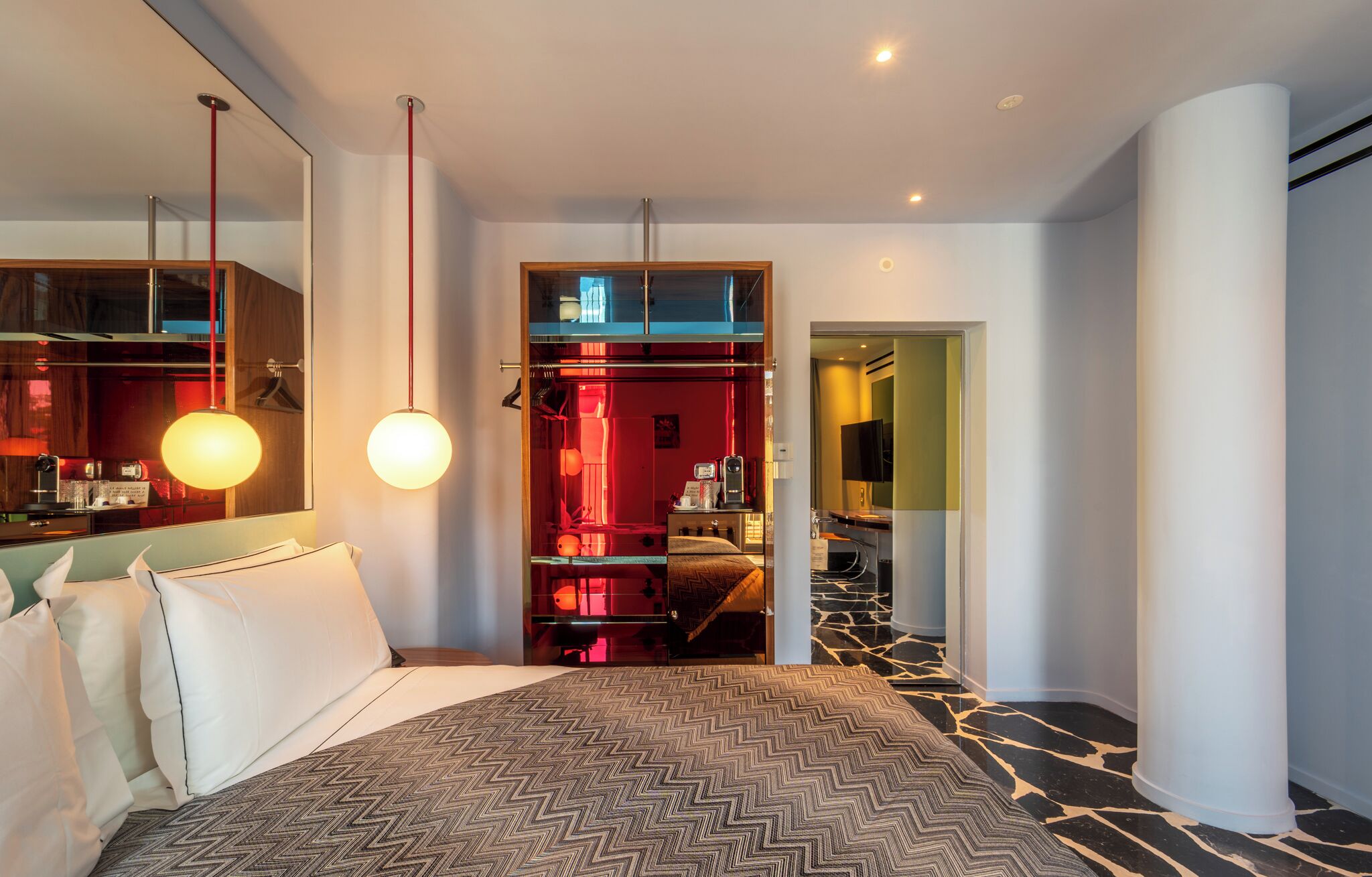 Habitación de hotel moderna con una cama grande, un vestidor, un espejo y un pasillo que conduce a un baño.