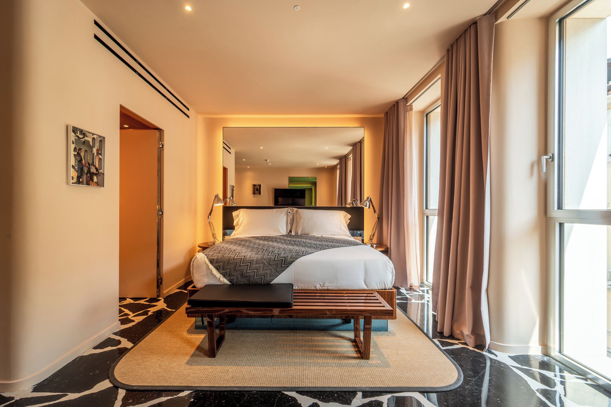 A modern hotel room with a large bed, a bench, and a view of the city through large windows.