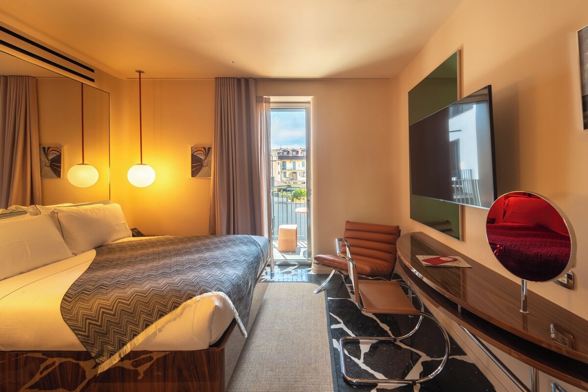 A hotel room with a bed, a desk, a chair, a TV, and a balcony with a view of buildings.