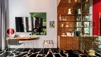 A modern hotel room with a large flat-screen TV, a wooden cabinet with glass doors displaying bottles and glasses, a round wooden desk with a chair, and a unique black and white patterned floor.