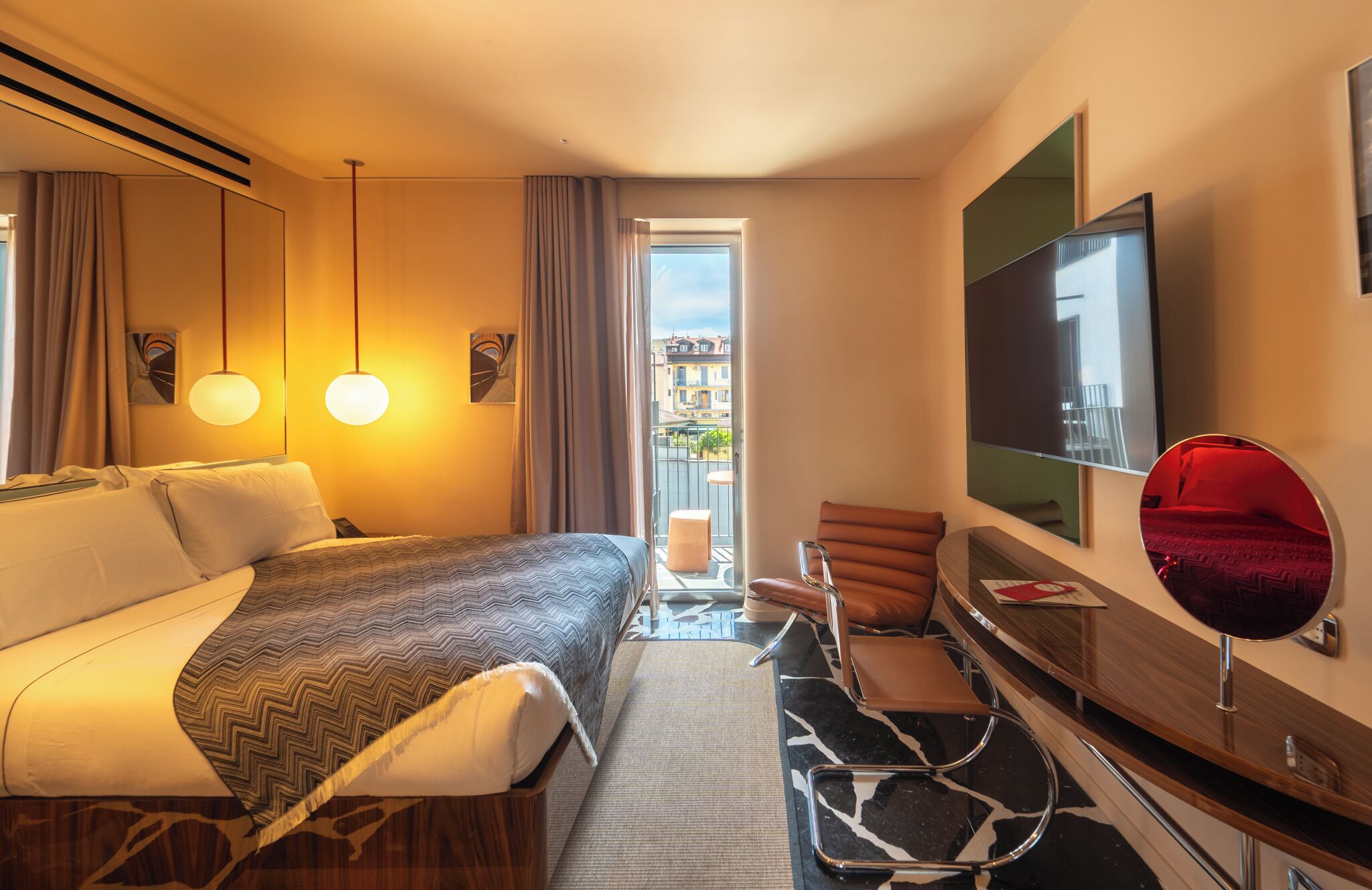 Ein Hotelzimmer mit Bett, Schreibtisch, Sessel, Fernseher und einem Balkon mit Blick auf Gebäude.
