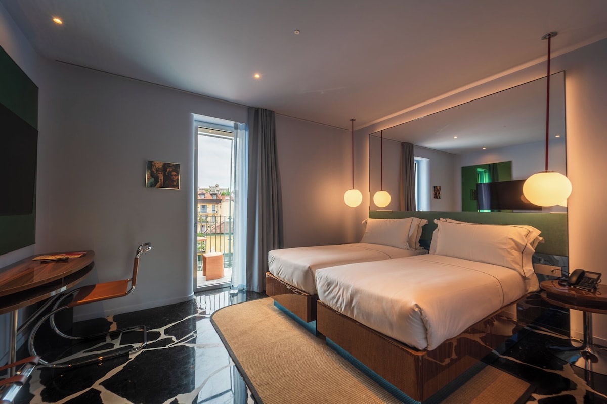 A modern hotel room with a large bed, a desk, a chair, a mirror, and a view of the outside.