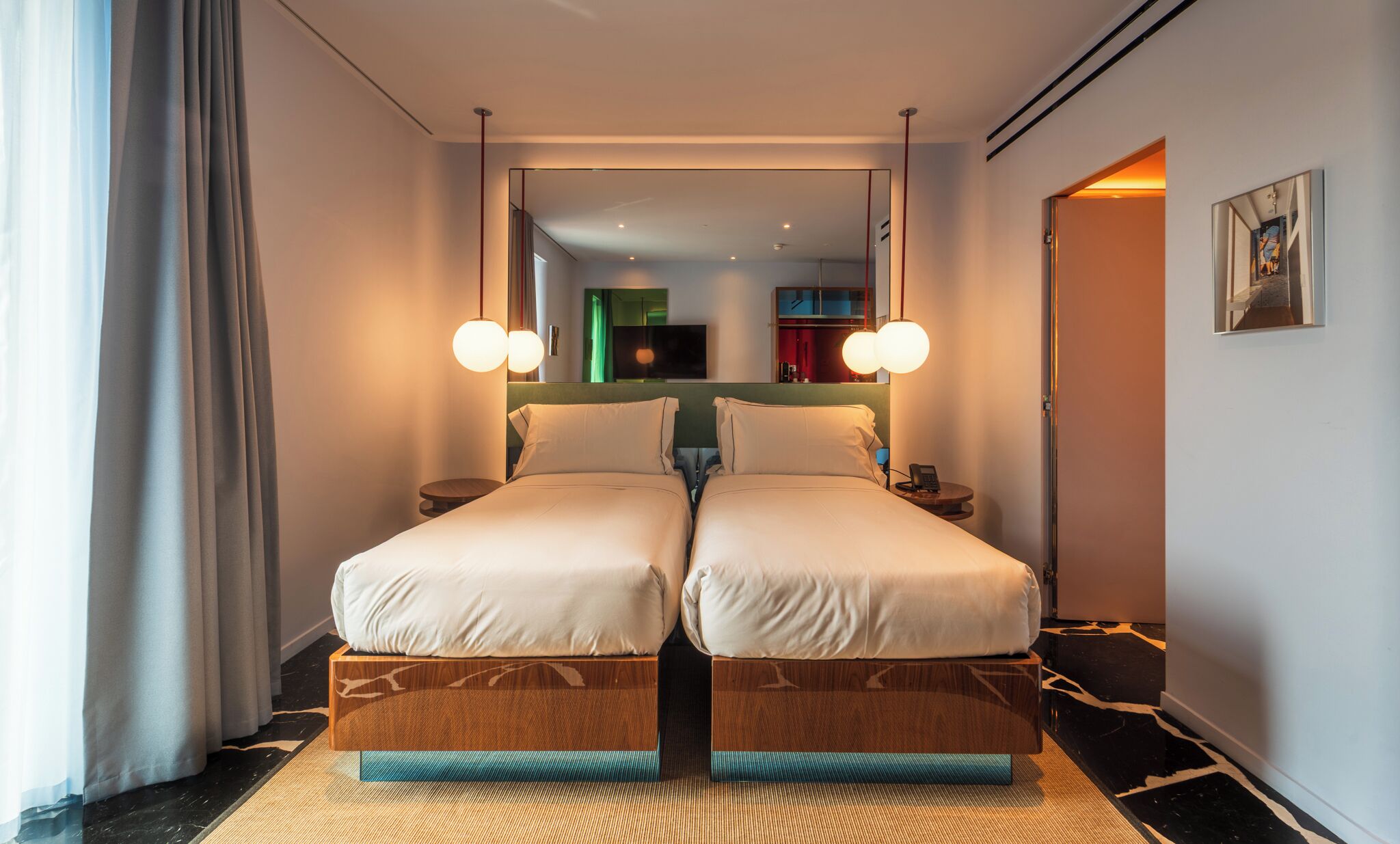 A double bed with a wooden frame and a mirror above it. There is a small bedside table with a lamp and a phone. The room has a black and white marble floor and a large window with curtains.