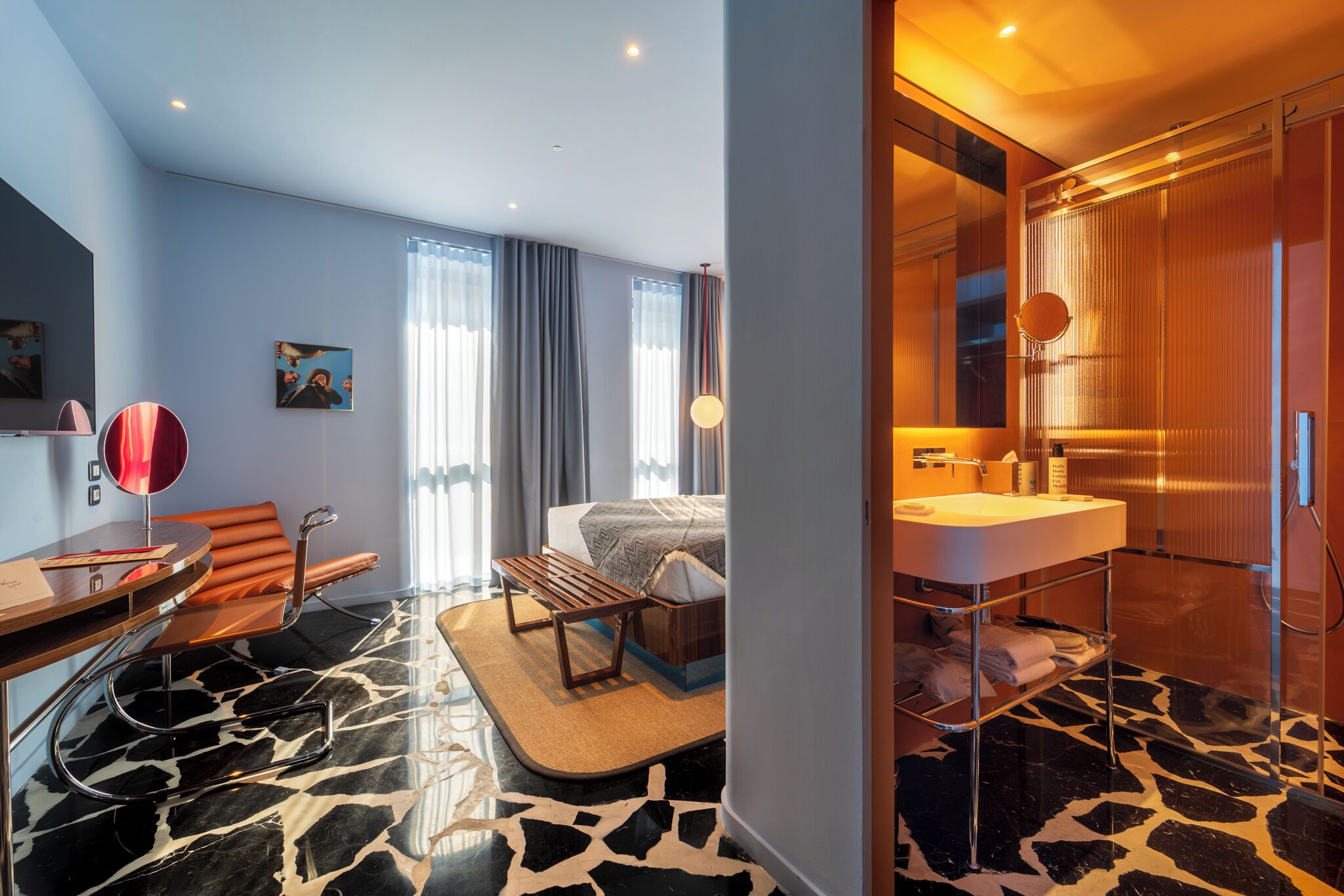 A modern hotel room with a bed, a desk, a chair, a bathroom with a mirror and sink, and a large window with curtains.
