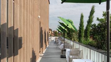 A covered outdoor dining area with tables and chairs, a wooden building on one side, and greenery on the other.