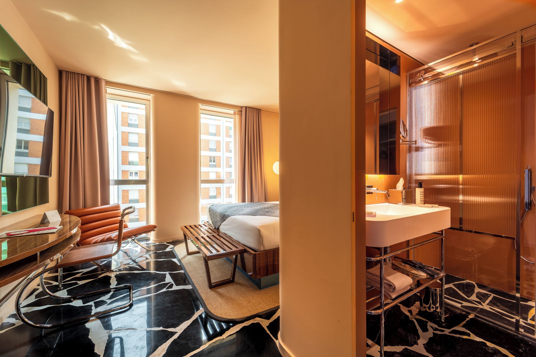 A hotel room with a bed, a desk with a chair, a large mirror, a bathroom with a sink and mirror, and a window with curtains.
