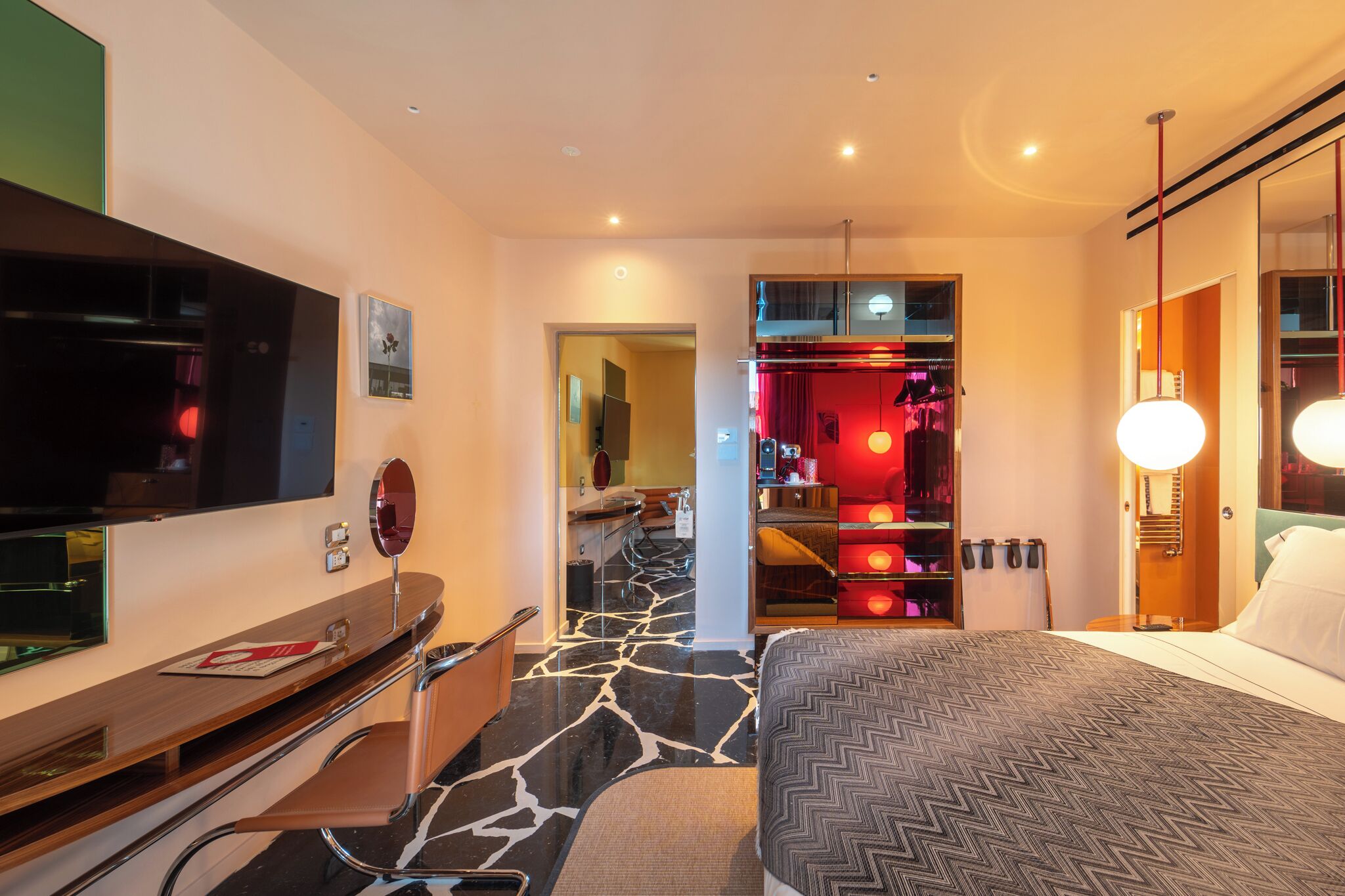 A modern hotel room with a large bed, a desk with a flat-screen TV, a vanity with a mirror, and a walk-in closet with red lighting.