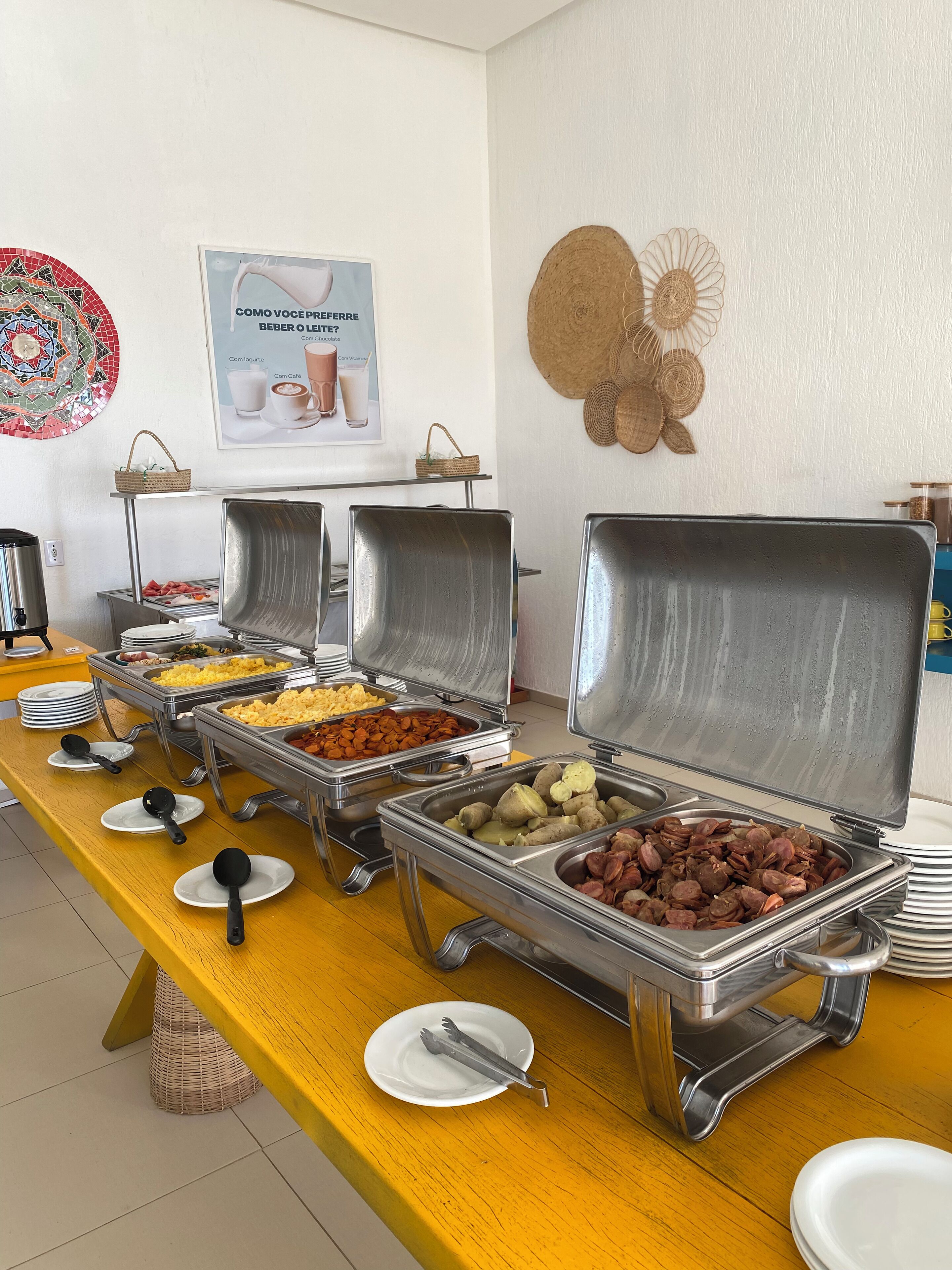 Montaje de bufé con diversos alimentos en recipientes metálicos, una mesa amarilla y una pared con un cartel y adornos.