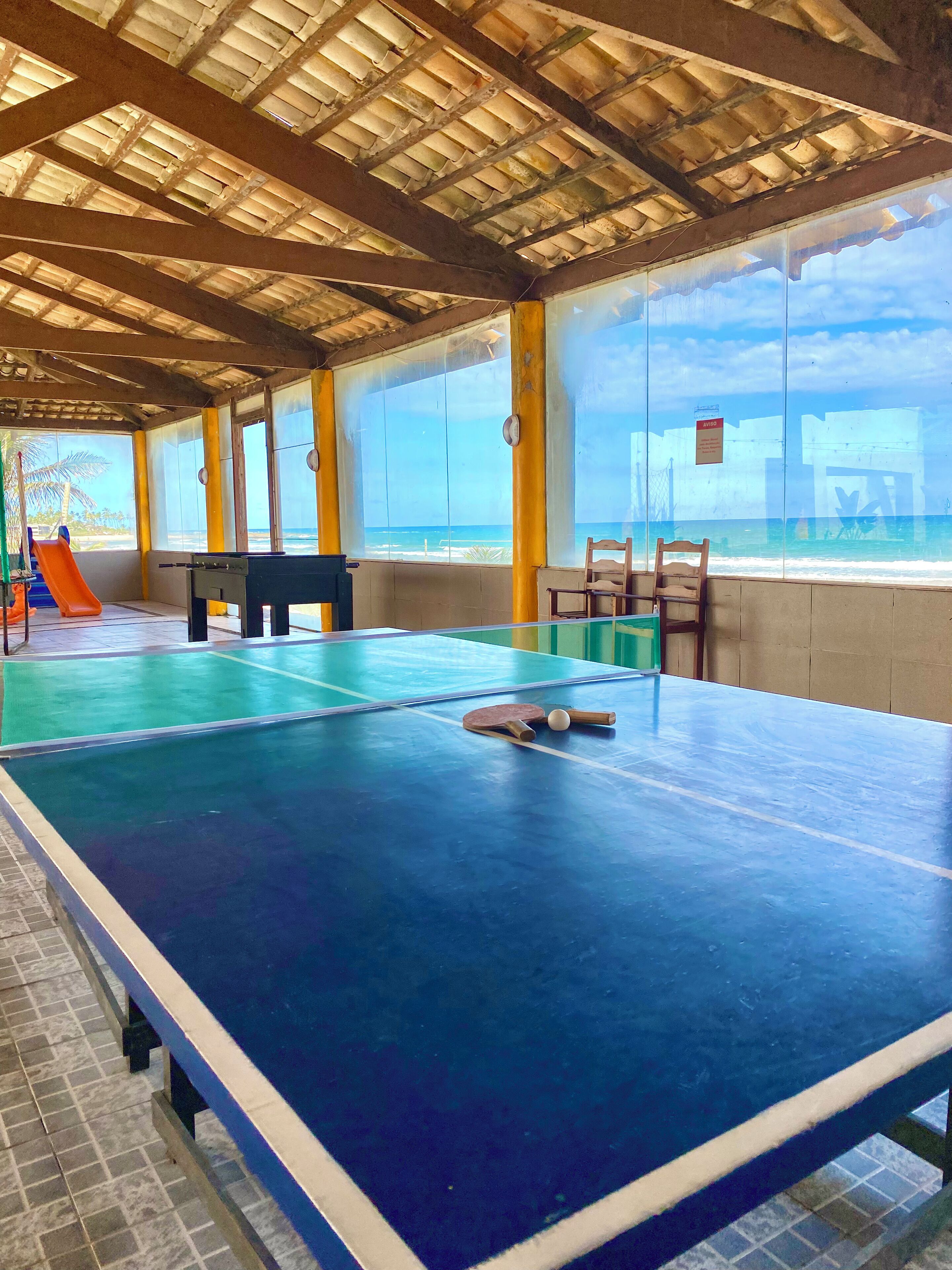 Uma mesa de pingue-pongue em ambiente interno, com vista para o oceano através de grandes janelas.