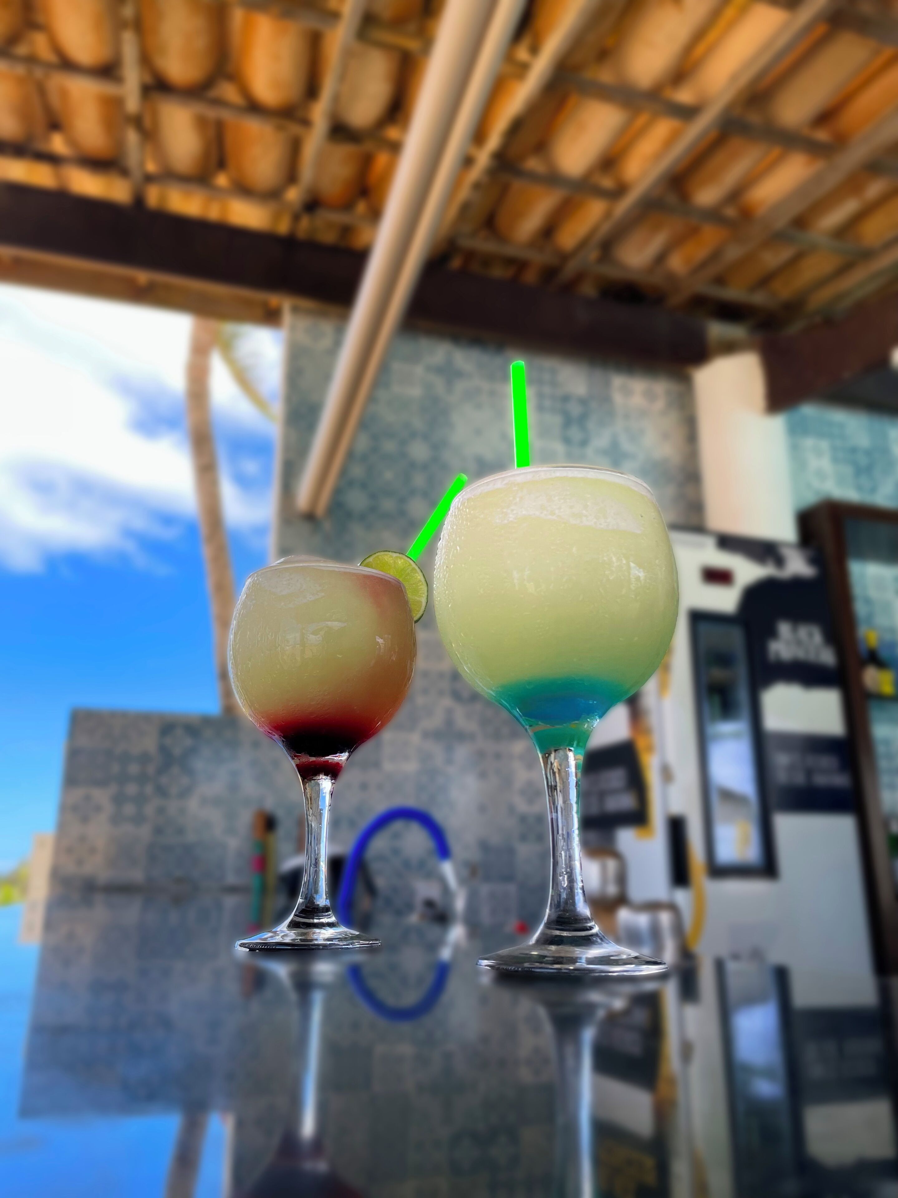 Two cocktails on a reflective surface with a tiled wall and a blue sky in the background.