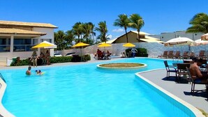 A swimming pool with lounge chairs and umbrellas, surrounded by palm trees and buildings. - Imperial Arembepe Beach Hotel (Camaçari)