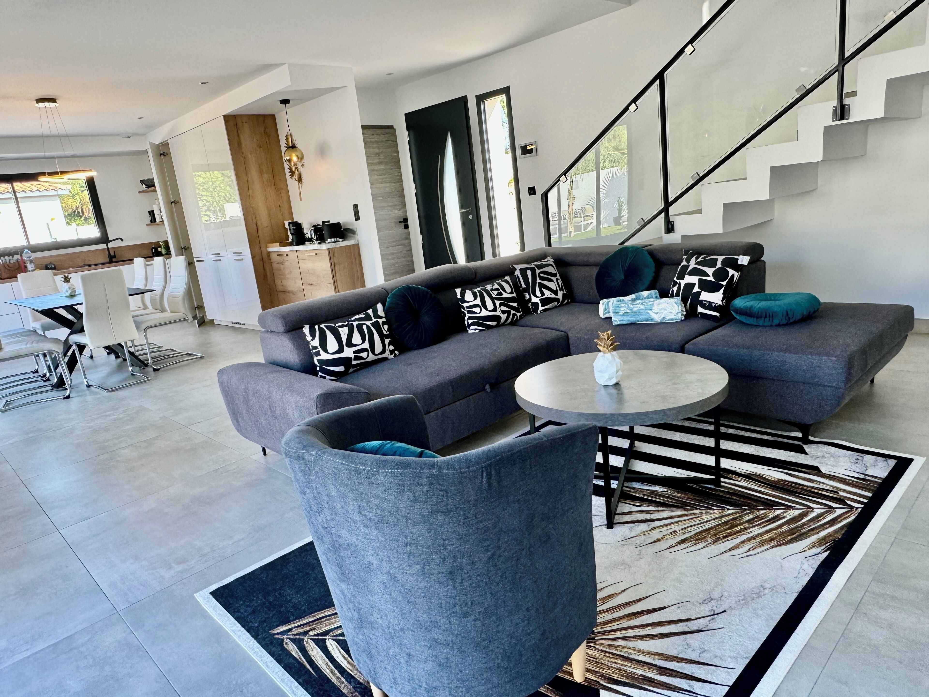 Un salon moderne avec un canapé d’angle gris, une table basse ronde et un escalier menant à un niveau supérieur.