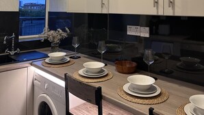 Uma cozinha moderna com máquina de lavar e secadora embutidas, mesa de jantar posta com tigelas brancas e taças de vinho, e vista da cidade pela janela.