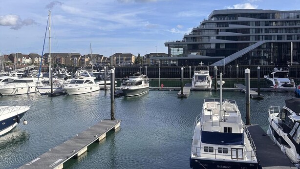 Marina com vários barcos atracados, um edifício moderno com fachada de vidro e céu claro.