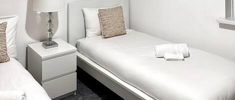 A single bed with a white mattress, a white pillow, and a beige throw pillow. There is a bedside table with a lamp and a small drawer. A white towel is placed on the bed.
