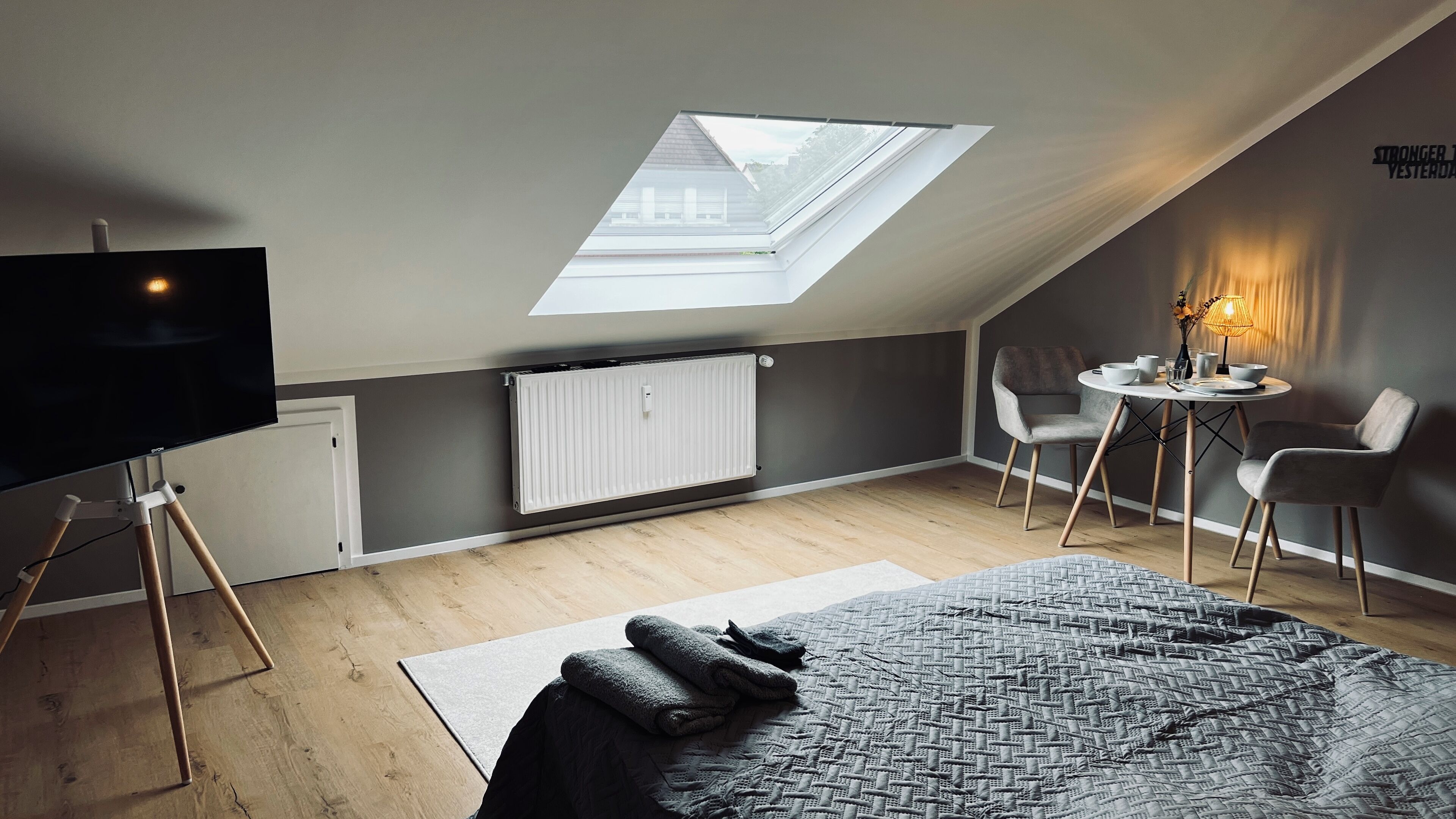 Une chambre moderne avec un puits de lumière, un téléviseur à écran plat, un radiateur, un lit recouvert d’une couverture à motifs et une petite table ronde avec des chaises.