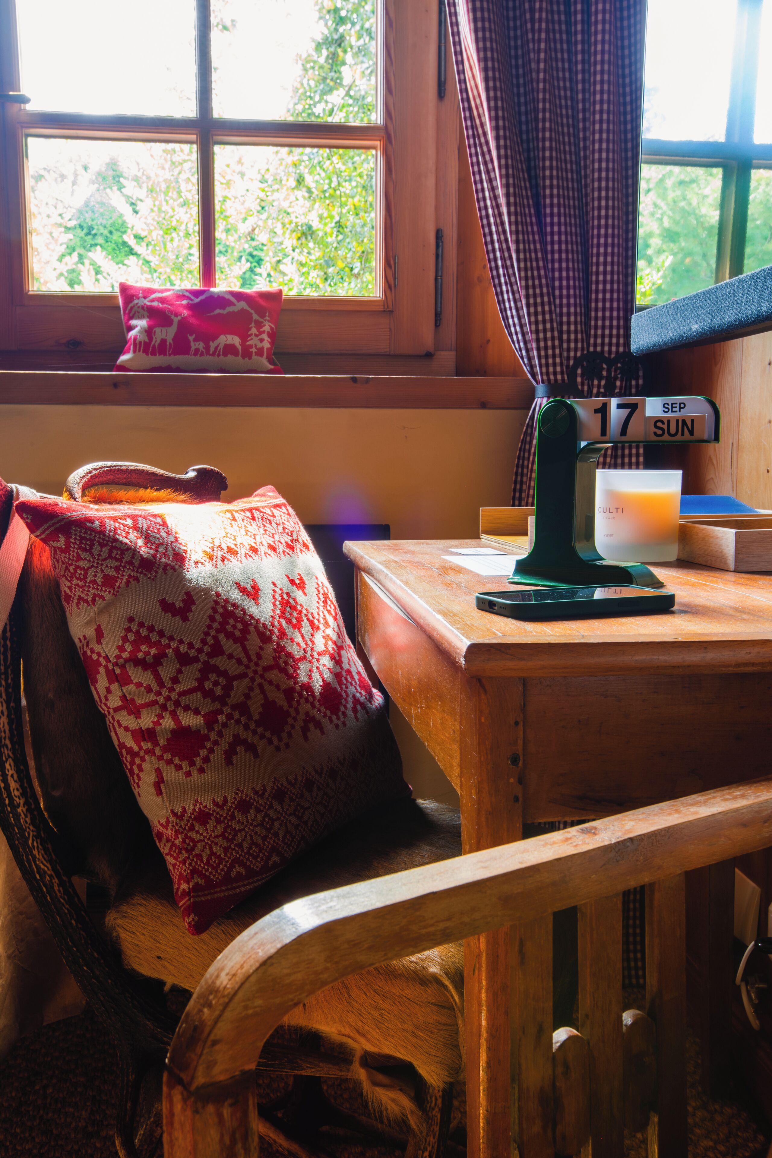 Une chaise en bois avec un coussin, une table en bois avec une bougie, un calendrier et une fenêtre avec des rideaux.