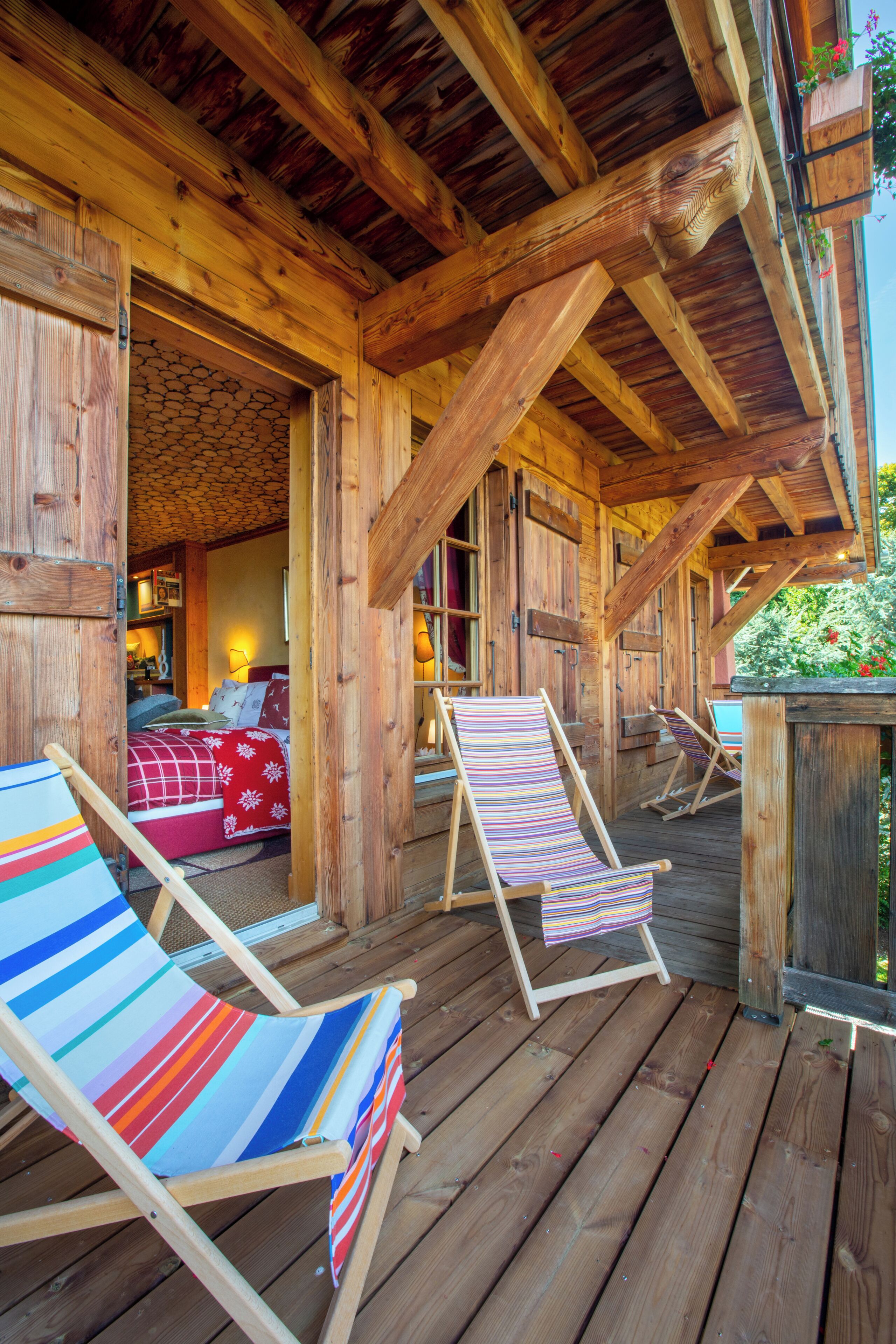 Una terraza de madera con dos sillones reclinables a rayas y una cama con manta de cuadros rojos y blancos.