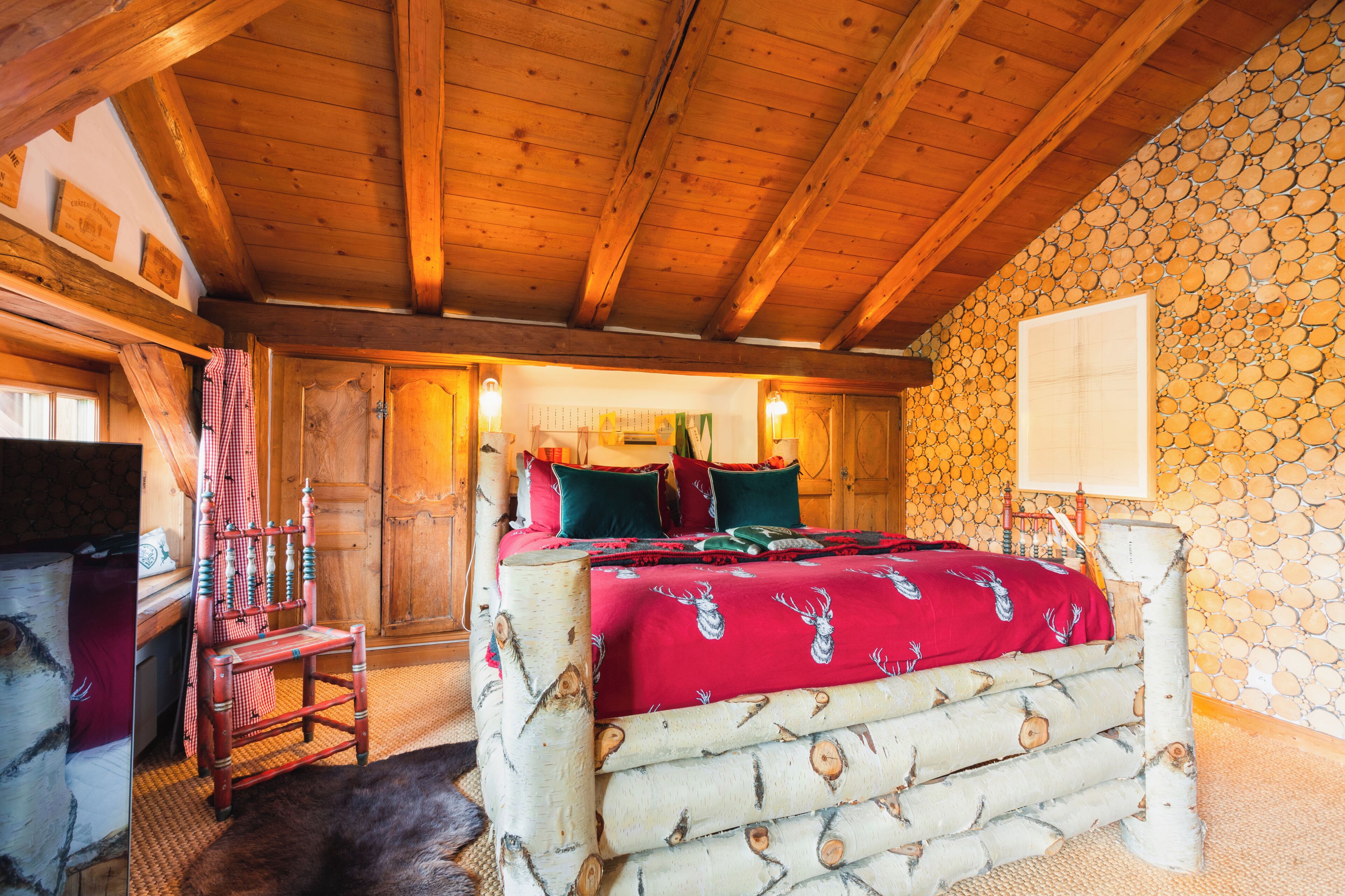 A wooden-framed bed with a red blanket, a wooden chair, and a stone wall.