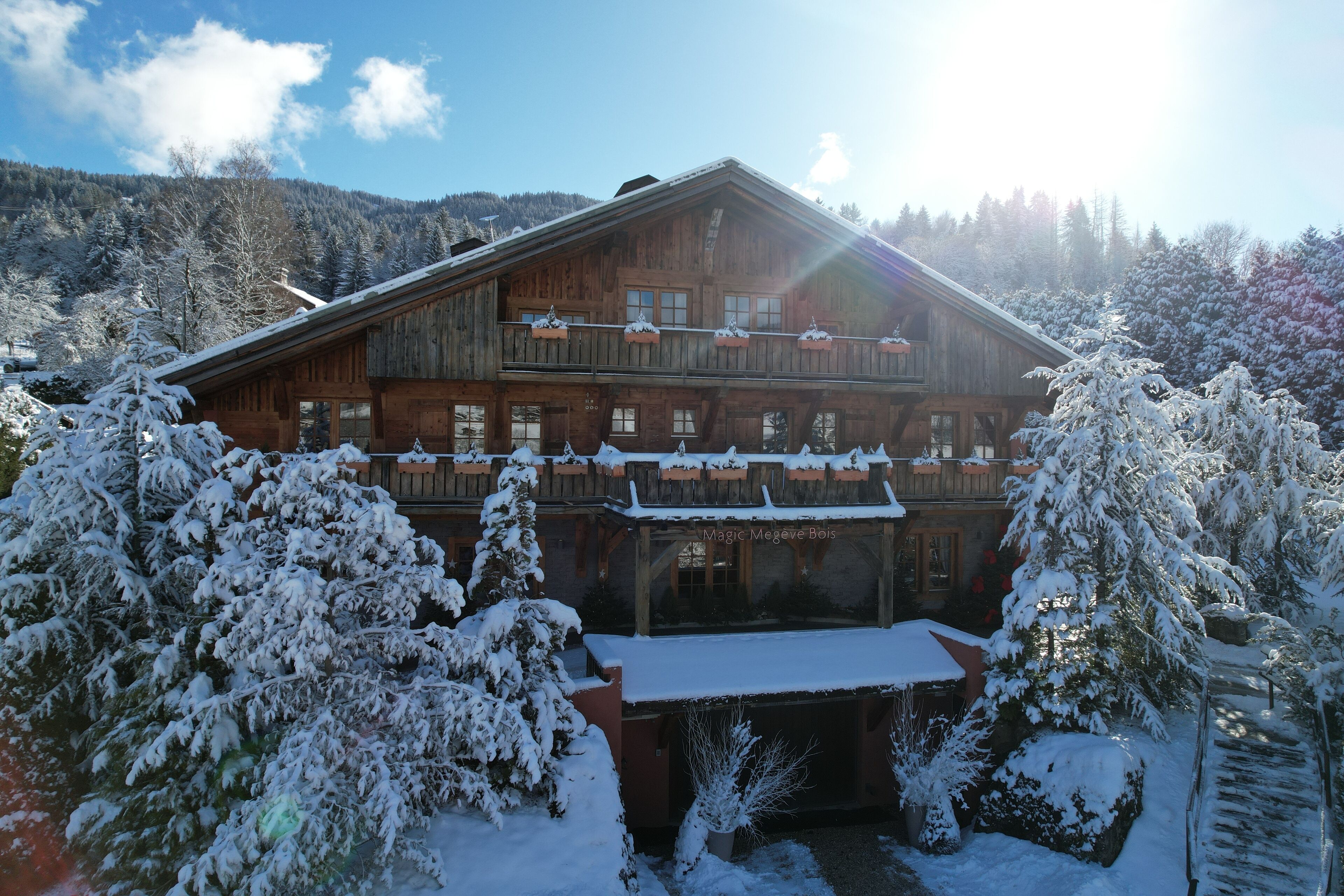 A wooden chalet with a balcony, surrounded by snow-covered trees and a mountainous backdrop.