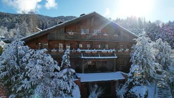 A wooden chalet with a balcony, surrounded by snow-covered trees and a mountainous backdrop.