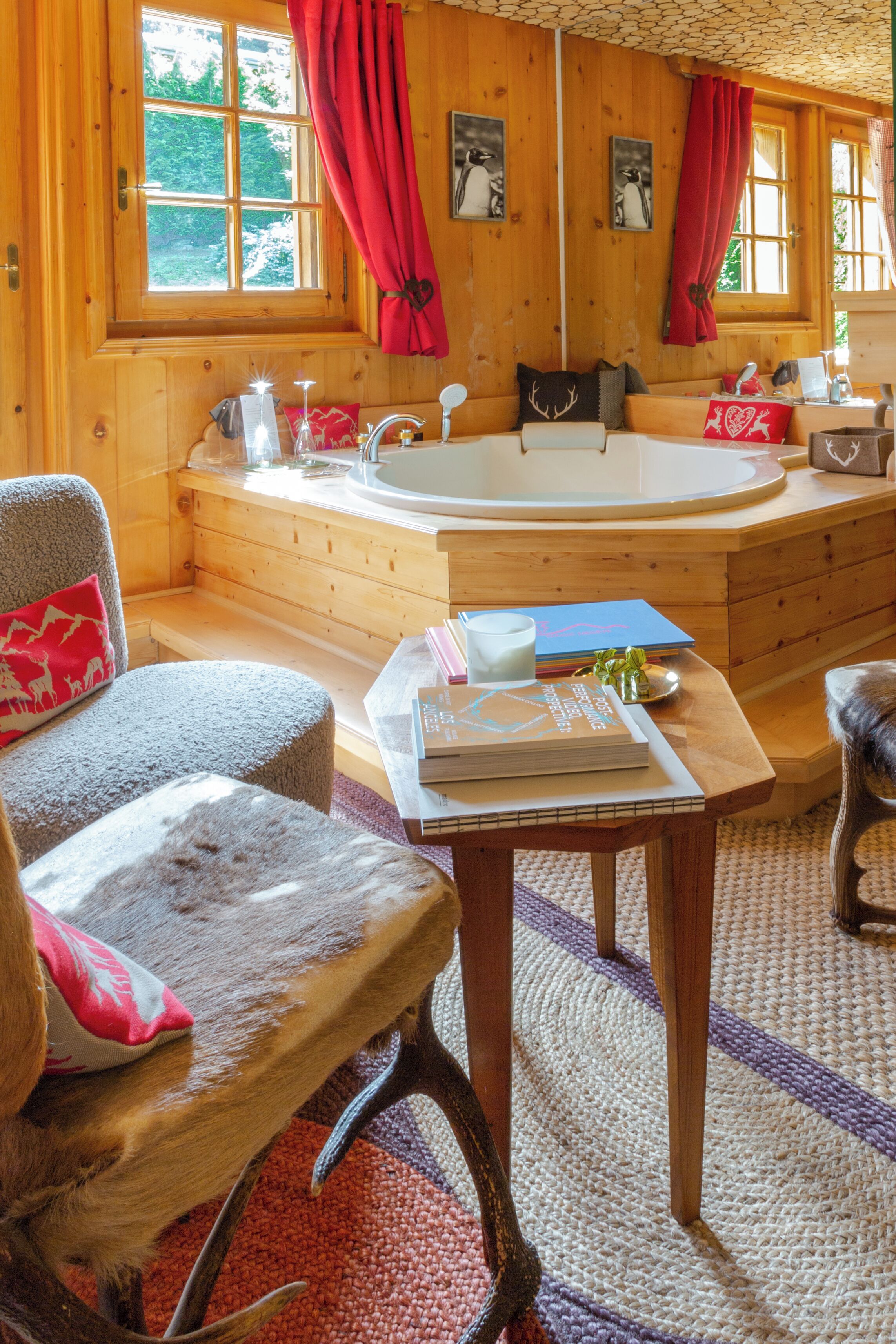 A wooden cabin interior with a wooden bathtub, a sofa, a table with books, and a window with red curtains.