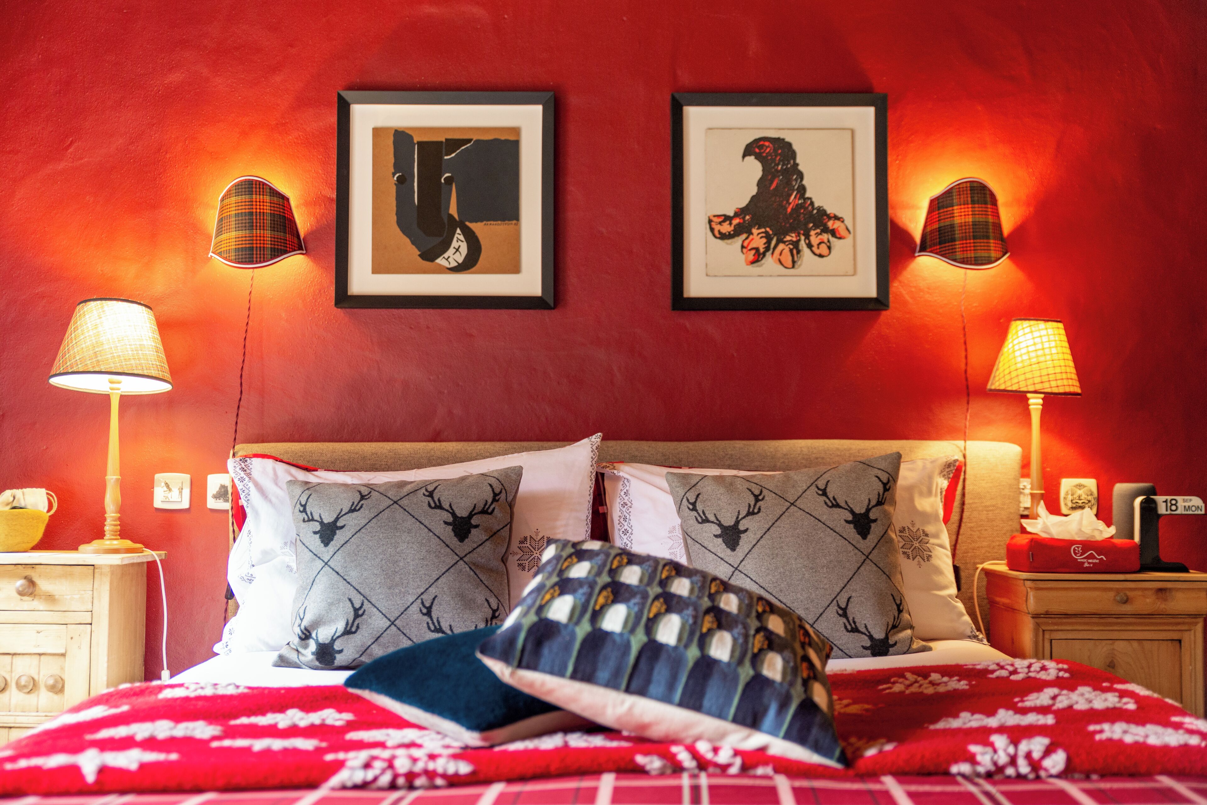 Hotelkamer met een rode muur, twee ingelijste afbeeldingen, een bed met gekleurde kussens, een rood-witte deken en twee bedlampen.