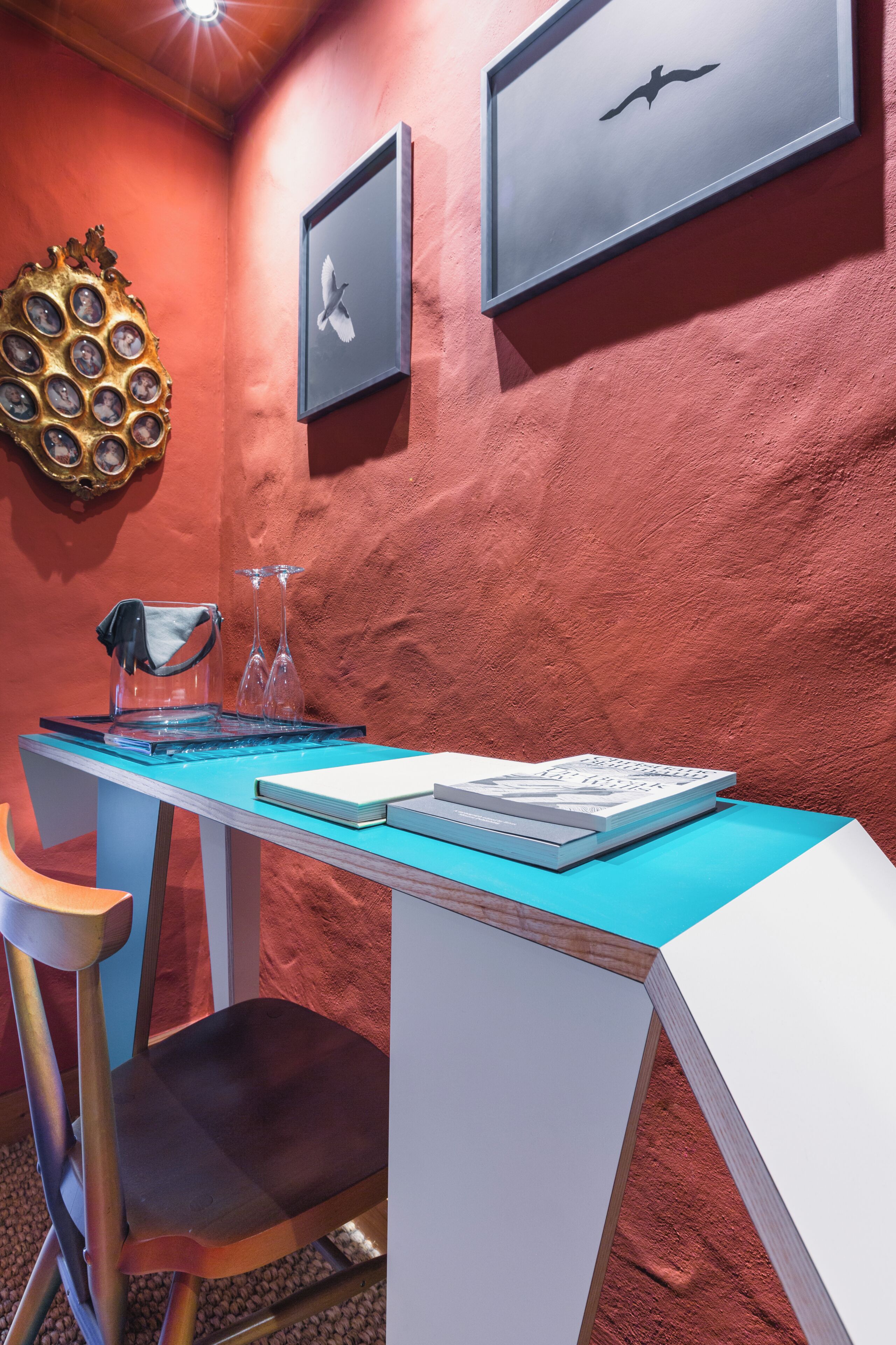 Una habitación con pared de textura roja, una mesa azul, una silla blanca y obras de arte enmarcadas.