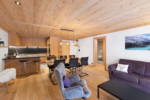 Um interior moderno com tetos de madeira, uma cozinha com uma ilha grande, uma sala de jantar com cadeiras e mesa, e uma sala de estar com sofá e um quadro.